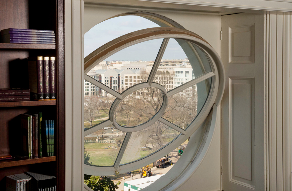 A circular library window framing the U.S. Capitol grounds and a federal office building, with rows of bound volumes on a dark wood shelf in the foreground.